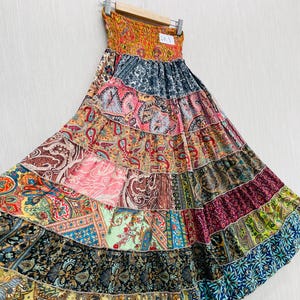 Puede incluir: Un vestido largo patchwork colorido con un corpiño sin tirantes y fruncido. La falda presenta un diseño escalonado con varios estampados de cachemira y florales en tonos rosa, azul, verde y amarillo. El vestido está colgado en una percha de madera.