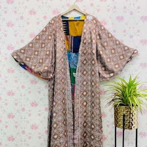 Könnte beinhalten: Langer, offen geschnittener Kimono-Mantel mit braunem und weißem geometrischem Muster. Der Mantel hat weite Ärmel und ein Patchwork-Design auf der Innenseite mit verschiedenen Farben. Er hängt an einem Holzbügel vor einem floralen Tapetenhintergrund.