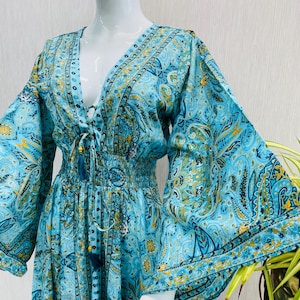 May include: A flowing, light blue dress with a paisley pattern in shades of blue and gold. The dress features a deep V-neckline, long, flared sleeves, and a cinched waist. A tassel detail hangs from the neckline.