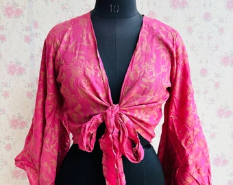 May include: A pink wrap top with long, flared sleeves and a tie closure. The top has a V-neck and a gold floral pattern. The garment is displayed on a black dress form against a floral wallpaper background.