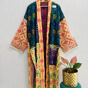 Könnte beinhalten: Ein langärmeliger Morgenmantel mit Patchwork-Design aus dunkelblauen, gelben und pfirsichfarbenen Stoffbahnen. Der Stoff ist mit aufwendigen Blumenstickereien in Pink, Gelb und Grün sowie kleinen reflektierenden Spiegelakzenten verziert.