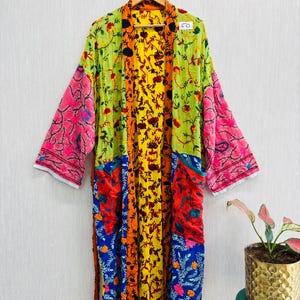 May include: A long-sleeved robe featuring a patchwork design with vibrant floral patterns in lime green, bright yellow, hot pink, deep blue, and orange. The garment includes a central yellow panel with dark floral motifs and a white tag labelled 50.