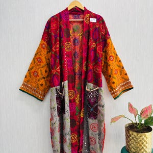 Può includere: Una vestaglia a maniche lunghe caratterizzata da un vivace design patchwork con pannelli rossi, arancioni e grigi. Il capo è decorato con intricati ricami floreali e paisley, piccoli specchietti riflettenti e due tasche frontali. È presente un'etichetta con il numero 34.