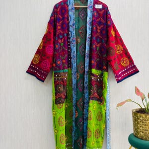 Könnte beinhalten: Ein langärmeliger Kimono-Bademantel mit einem lebendigen Patchwork aus magentafarbenem, hellgrünem und dunkelgrünem Stoff. Das Kleidungsstück ist mit aufwendigen, mehrfarbigen Blumenstickereien, kleinen reflektierenden Spiegelakzenten und einer hellblauen Borte verziert.