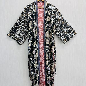 Puede incluir: Larga bata kimono abierta por delante, con un estampado floral en negro, marrón y blanco. El forro interior es rosa y rojo. La bata está colgada de una percha de madera.