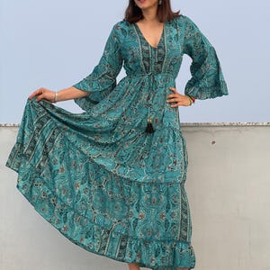 May include: A teal maxi dress with a paisley pattern. The dress features long, flared sleeves, a V-neck, and a drawstring waist. The skirt has a tiered design with a ruffled hem. The dress is worn with clear heeled sandals.