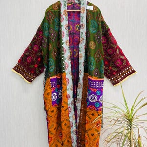 Könnte beinhalten: Ein langärmeliger Kimono-Bademantel mit Patchwork-Design in Olivgrün, Dunkelviolett und leuchtendem Orange. Das Kleidungsstück ist mit aufwendigen, mehrfarbigen Blumenstickereien, kleinen runden Spiegeln und einer hellblauen Bordüre verziert.