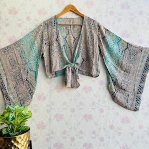 Puede incluir: Un top corto con nudo delantero y mangas largas y anchas. El top está hecho de una tela ligera con un estampado paisley en tonos azules, marrones y blancos.