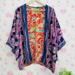 May include: A patterned kimono jacket in navy blue, pink, and white paisley. The lining is red with a floral design. The jacket has a blue trim and is displayed on a wooden hanger. The garment is a stylish outerwear piece.