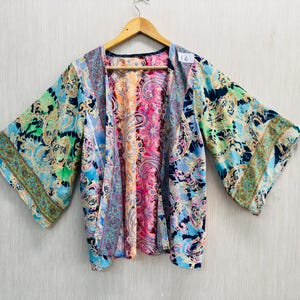 Puede incluir: Una colorida parte superior estilo kimono con un vibrante estampado de cachemira en tonos de azul, verde, rosa y naranja. La parte superior tiene mangas anchas y está colgada de una percha de madera. La prenda es de tejido ligero y fluido.