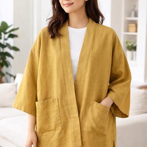 Pode incluir: Casaco kimono de linho amarelo mostarda com dois bolsos frontais grandes. O casaco tem um corte folgado e mangas três quartos. A modelo está a usar uma t-shirt branca e calças de ganga azuis. O casaco é aberto na frente.