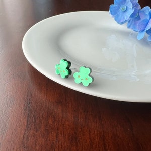 Pode incluir: Um par de brincos verdes em forma de flor com contornos pretos e rostos pintados a ouro. Os brincos est&atilde;o num prato branco.
