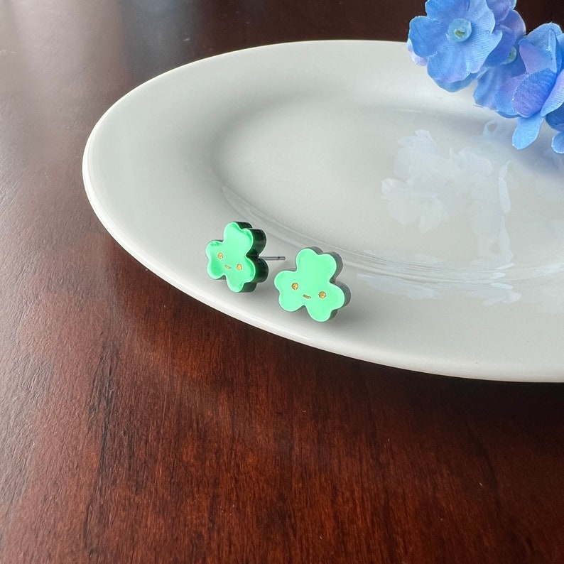 Pode incluir: Um par de brincos verdes em forma de flor com contornos pretos e rostos pintados a ouro. Os brincos est&atilde;o num prato branco.