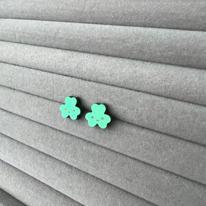 Pode incluir: Um par de brincos verdes em forma de trevo com contorno preto e um rosto sorridente dourado em cada brinco.