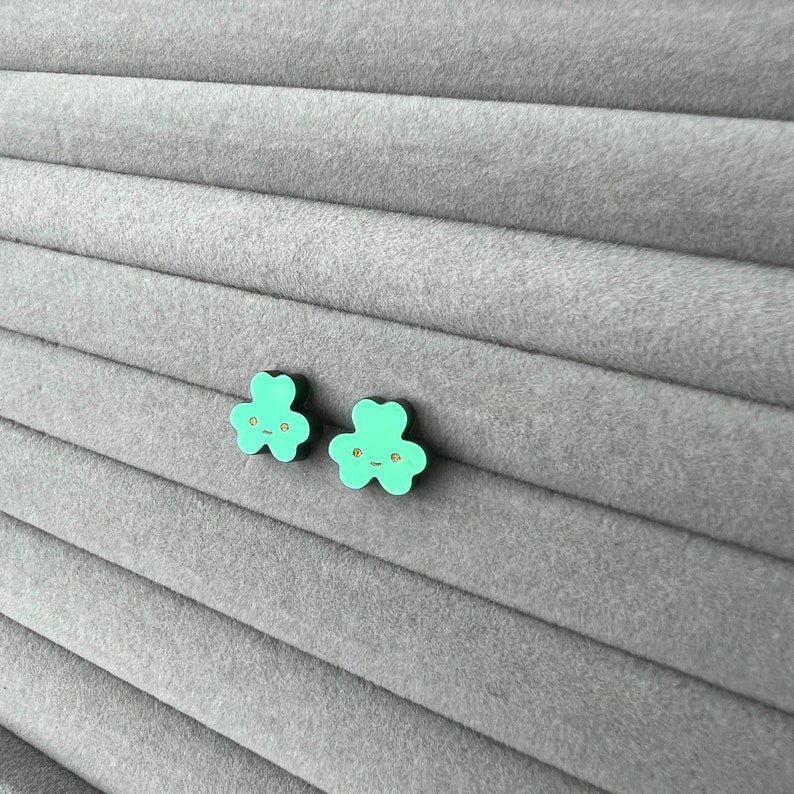 Pode incluir: Um par de brincos verdes em forma de trevo com contorno preto e um rosto sorridente dourado em cada brinco.