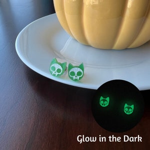 May include: A pair of green cat skull earrings with white details. The earrings glow in the dark, as shown in the image.