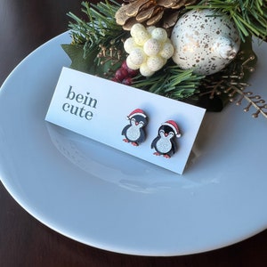 May include: A pair of black and white penguin earrings with red Santa hats. The earrings are on a white card with the text "bein cute".