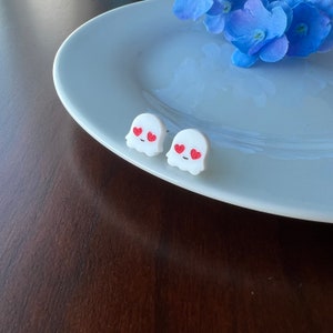 May include: A pair of white ghost-shaped earrings with red heart eyes. The earrings are on a white plate.