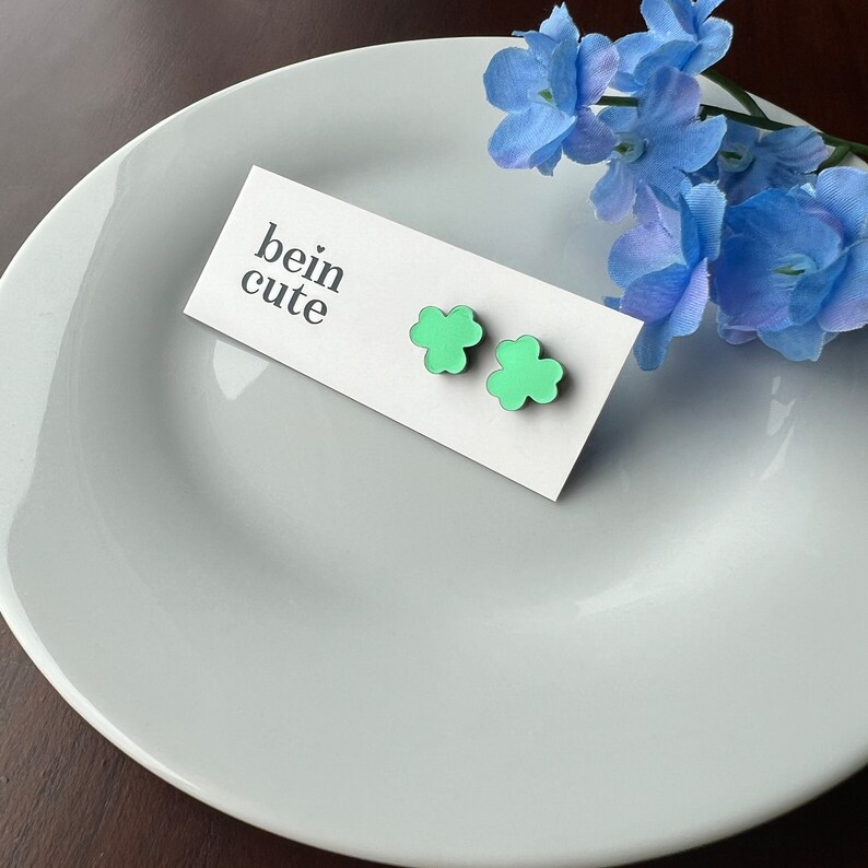 Pode incluir: Um par de brincos verdes em forma de trevo em um cart&atilde;o branco com o texto "bein cute".