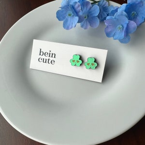 Pode incluir: Um par de brincos de trevo verde com detalhes em cora&ccedil;&atilde;o dourado. Os brincos est&atilde;o em um cart&atilde;o branco com o texto "bein cute".