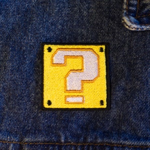 Puede incluir: Un bloque amarillo con forma de signo de interrogación con un signo de interrogación blanco en su interior. El bloque está bordado en una chaqueta vaquera azul.