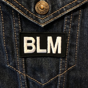 May include: A black patch with white letters that spell out "BLM" is sewn onto a blue denim jacket.