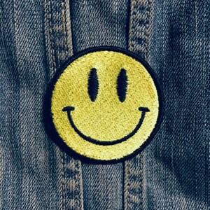 Op de afbeelding: Een gele smiley-patch met een zwarte omtrek, genaaid op een blauw denim jack.