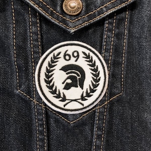 Puede incluir: Un parche bordado blanco y negro con un casco espartano y una corona de laurel. El parche tiene el número "69" en el centro. El parche está cosido en una chaqueta vaquera oscura.