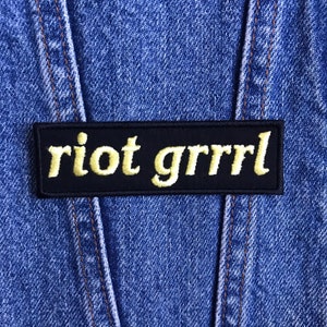 May include: A black rectangular patch with the words "riot grrrl" embroidered in yellow script. The patch is set against a blue denim background, with visible stitching.