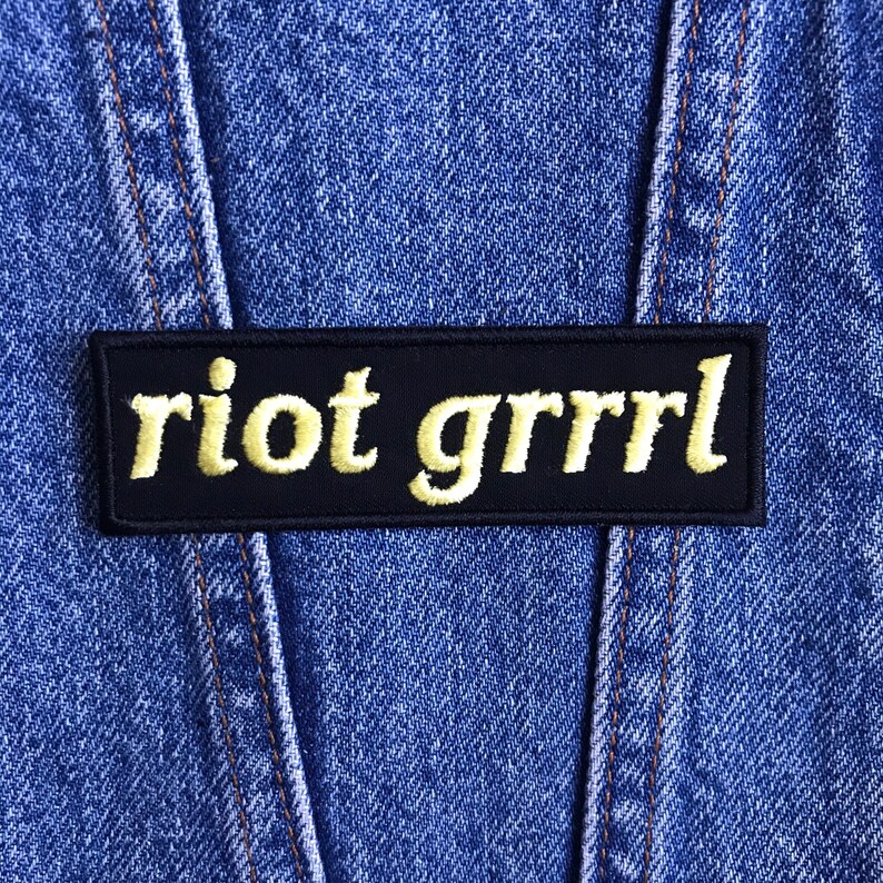 May include: A black rectangular patch with the words "riot grrrl" embroidered in yellow script. The patch is set against a blue denim background, with visible stitching.