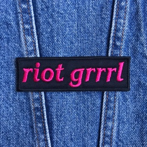 May include: A rectangular black patch with the words "riot grrrl" embroidered in bright pink thread. The patch is affixed to a blue denim jacket, with visible seams and stitching.