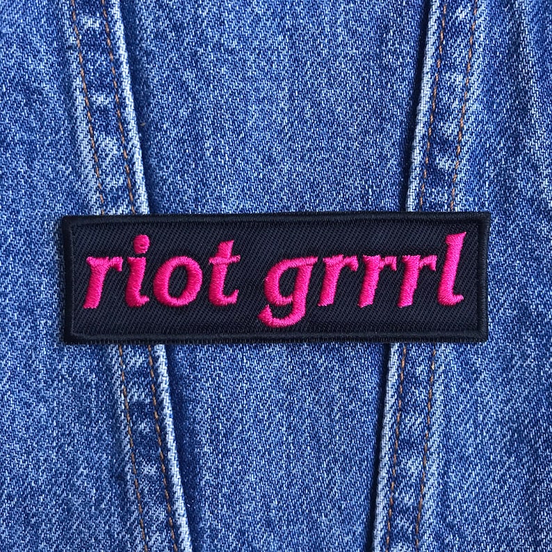 May include: A rectangular black patch with the words "riot grrrl" embroidered in bright pink thread. The patch is affixed to a blue denim jacket, with visible seams and stitching.