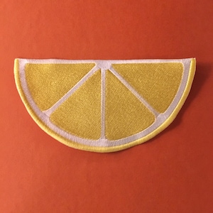 May include: A yellow lemon wedge embroidered patch with white stitching. The patch is shaped like a slice of lemon and has a white outline.