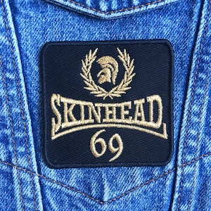 Op de afbeelding: Een zwarte stoffen patch met gouden borduurwerk. De patch heeft een lauwerkrans met een Spartaanse helm erin, het woord "SKINHEAD" in een gebogen lettertype en het nummer "69" eronder.
