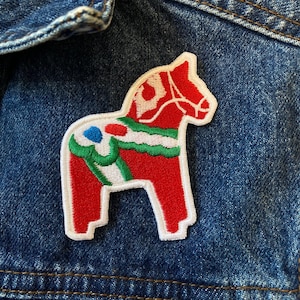 Puede incluir: Un parche bordado de un caballo Dala rojo y blanco con detalles verdes y azules. El parche está cosido en una chaqueta vaquera azul.