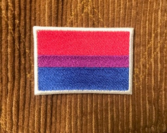 May include: A white-bordered patch with a bisexual pride flag design. The flag has three horizontal stripes: pink, purple, and blue.