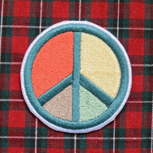 Op de afbeelding: Geborduurde vredesteken patch met oranje, gele, bruine en groene segmenten, omlijnd met turquoise en witte stiksels. De patch is geplaatst op een rood en groen geruit stoffen achtergrond.