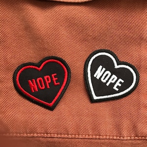 May include: Two heart-shaped patches with the word "NOPE". One patch is black with a red border and red text. The other is black with a white border and white text. The patches are on a brown fabric.