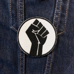 Pode incluir: Um remendo circular branco com um punho negro levantado no centro. O remendo é costurado em um tecido de sarja azul escuro.