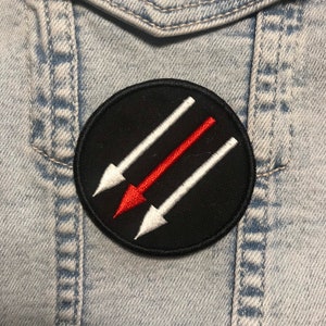 May include: A round, black embroidered patch featuring three arrows. Two white arrows flank a central red arrow, all pointing downwards. The patch is affixed to a light blue denim jacket.