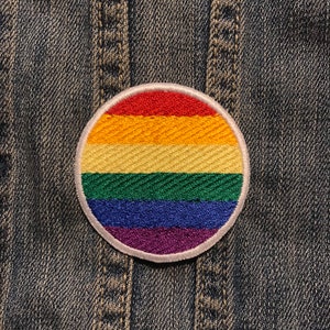 May include: A round, embroidered patch featuring the rainbow colors of the Pride flag: red, orange, yellow, green, blue, and purple. The patch has a white border and is set against a denim background.