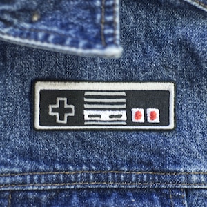 Puede incluir: Un parche bordado en blanco y negro de un mando de videojuego clásico con botones rojos. El parche está cosido en una chaqueta vaquera azul.
