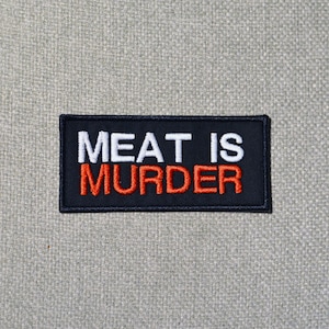 Może przedstawiać: Czarna prostokątna naszywka z wyhaftowanym napisem "MEAT IS MURDER" w kolorze białym i czerwonym. Naszywka ma czarną obwódkę i jest umieszczona na jasnoszarym, teksturowanym tle.