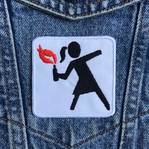 May include: White patch with a black silhouette of a person throwing a Molotov cocktail. The person has two pigtails and is wearing a short skirt. The Molotov cocktail is red and orange.