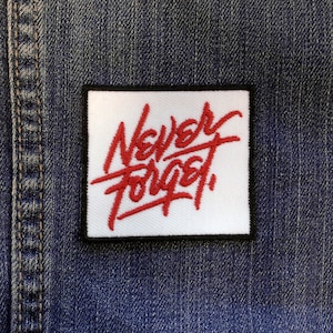 May include: White square patch with black border, featuring red embroidered text that reads "Never Forget".