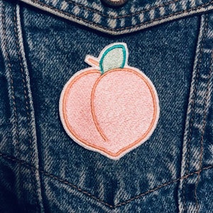Könnte beinhalten: Ein gestickter Pfirsich-Patch in Pink mit einem grünen Blatt auf einer blauen Jeansjacke.
