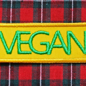 Puede incluir: Un parche rectangular amarillo con la palabra "VEGAN" bordada en verde sobre un fondo de cuadros rojos y negros.