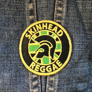 Può includere: Una toppa rotonda con le parole "SKINHEAD REGGAE" in lettere gialle. La toppa presenta un disegno nero e verde di un elmo e un motivo a scacchiera, su uno sfondo di denim.