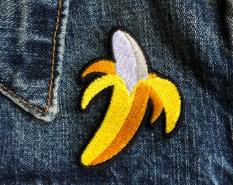 Puede incluir: Un parche bordado de plátano amarillo con cáscara blanca y contorno negro. El parche está cosido en una chaqueta vaquera azul.