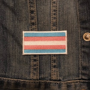 Puede incluir: Un parche rectangular bordado con la bandera del orgullo transgénero, con franjas horizontales en azul claro, rosa y blanco. El parche está bordeado con un contorno blanco y está fijado a una chaqueta vaquera oscura.